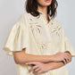 Woman wearing a cream short-sleeve embroidered blouse with eyelet details and tie front