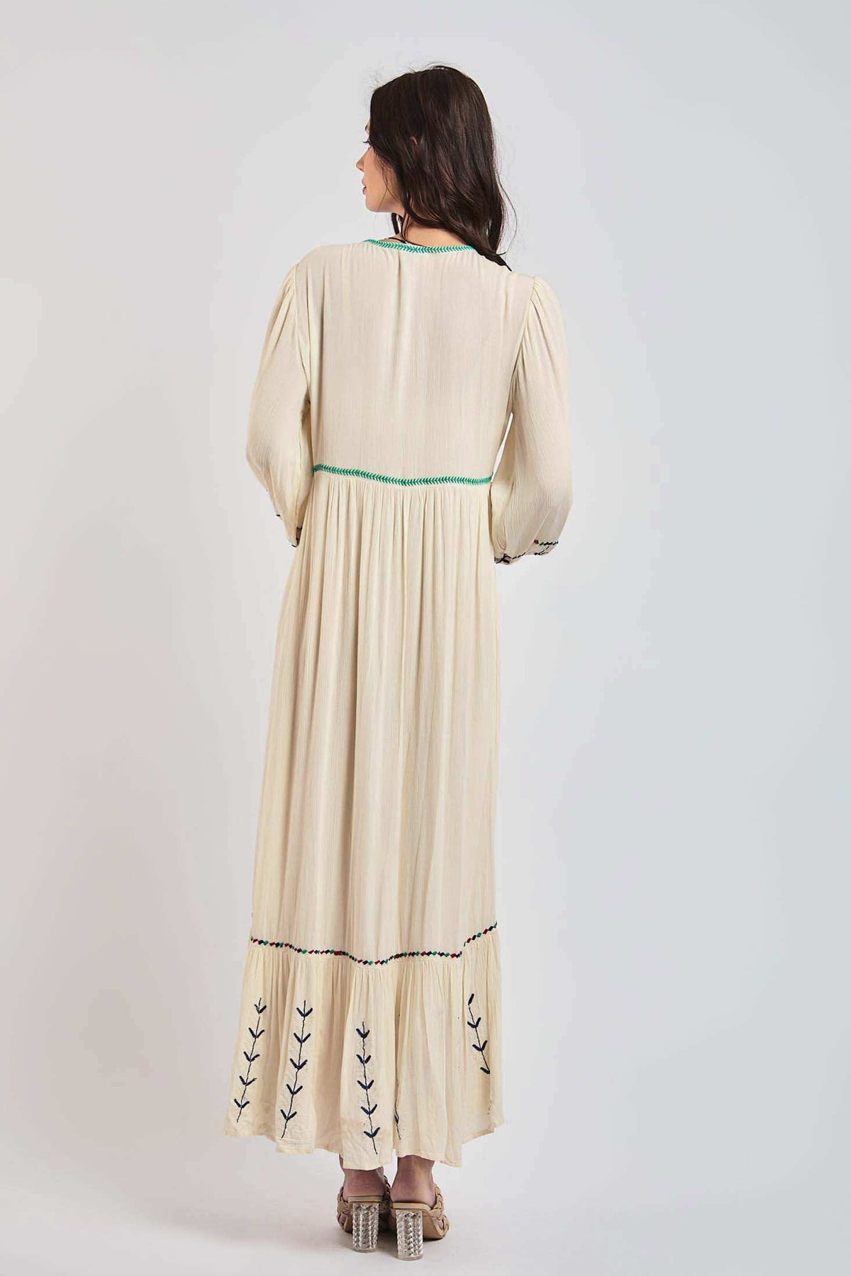 Back view of woman wearing long cream dress with embroidered hem and green accent stitching