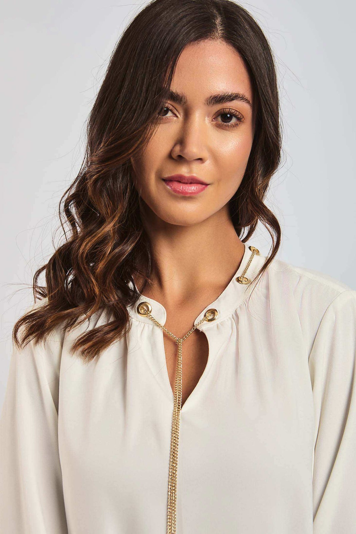 Woman with wavy hair wearing a white blouse with gold button details and chain necklace
