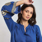 Woman wearing blue embroidered long sleeve blouse with gold details, posing indoors