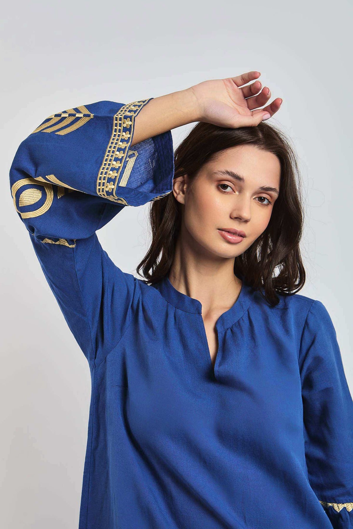 Woman wearing blue embroidered long sleeve blouse with gold details, posing indoors