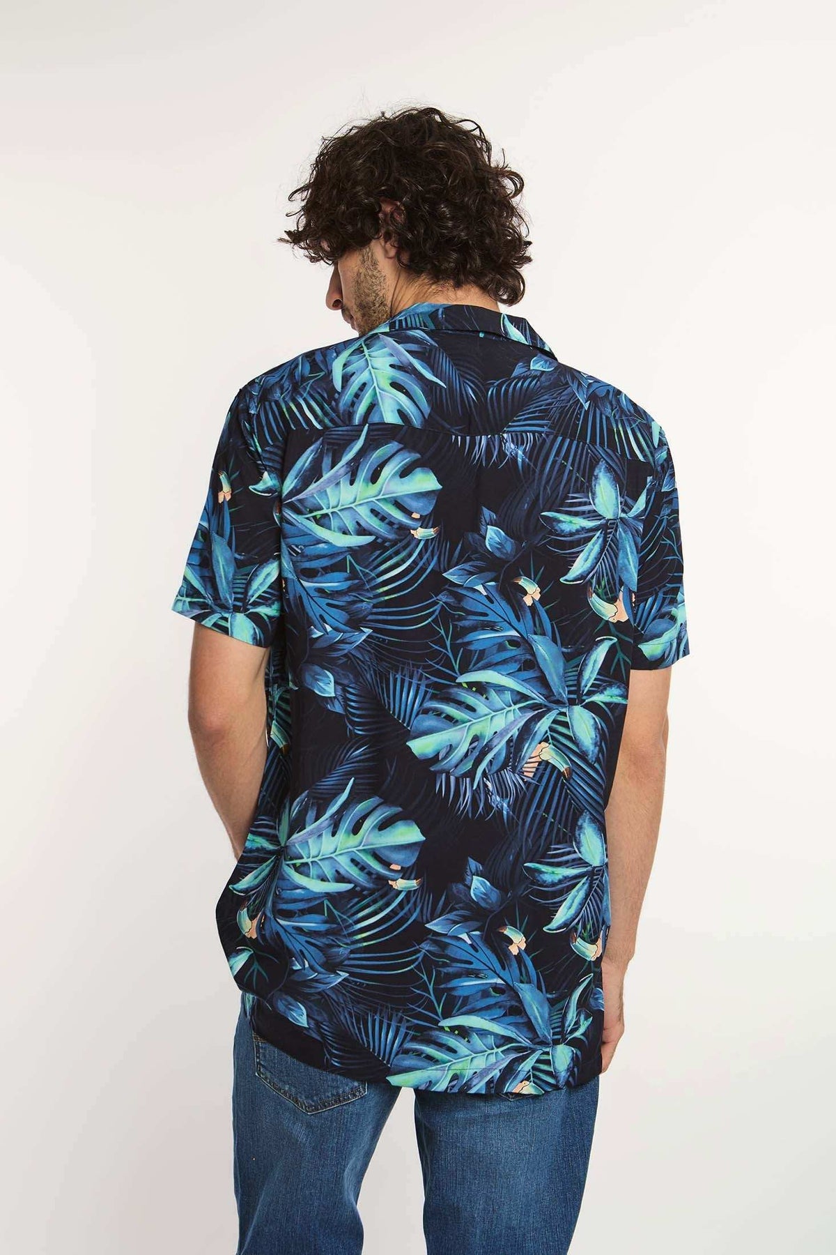 Man wearing blue tropical leaf print short sleeve shirt and denim jeans, back view