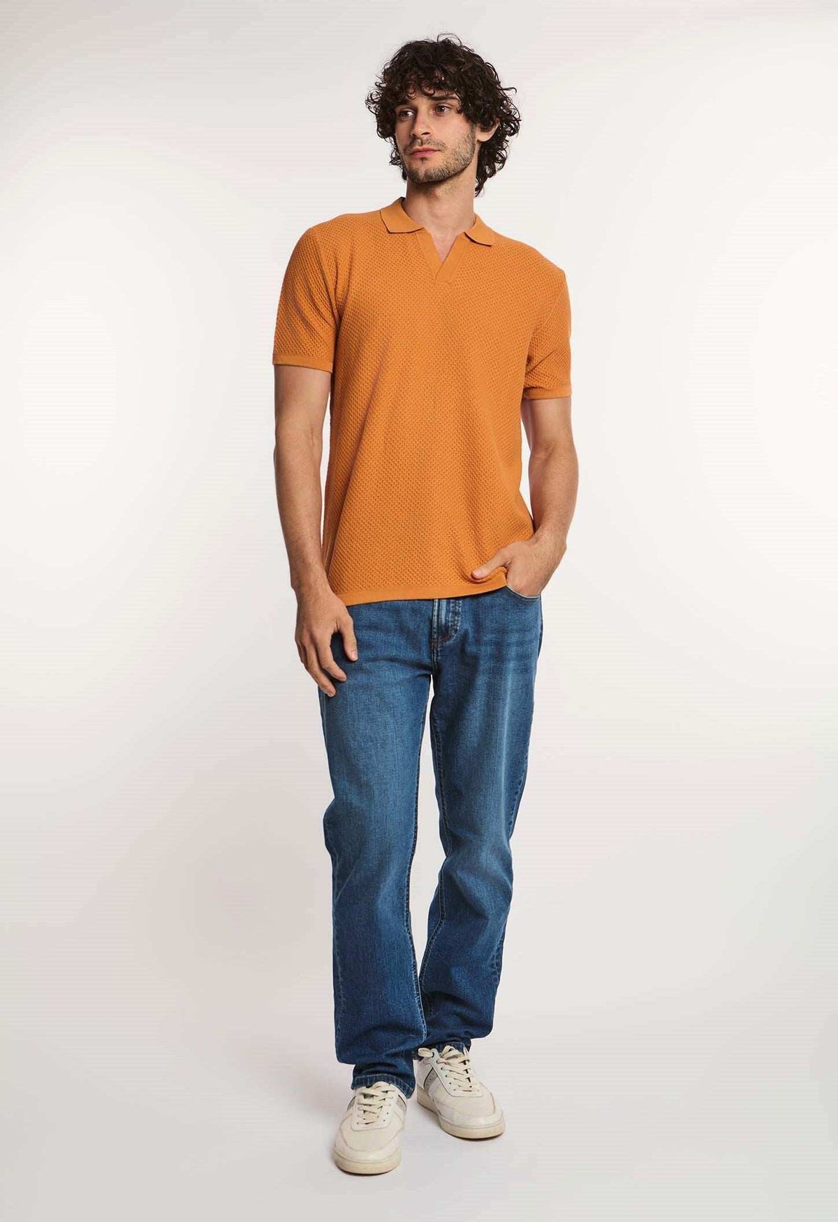 Man in an orange textured polo shirt, blue jeans, and white sneakers standing casually