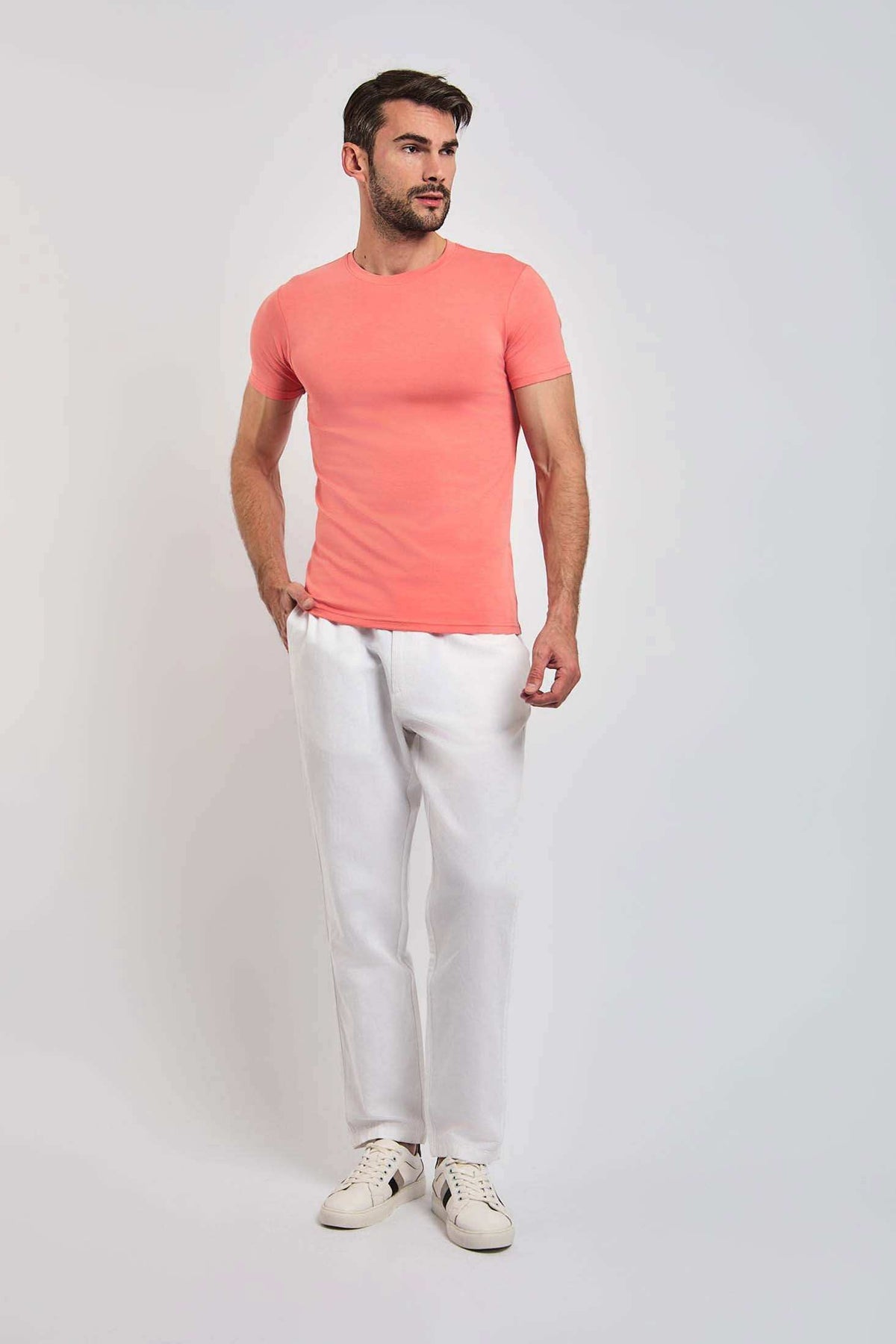 Man wearing a coral short-sleeve t-shirt, white pants, and white sneakers on plain background