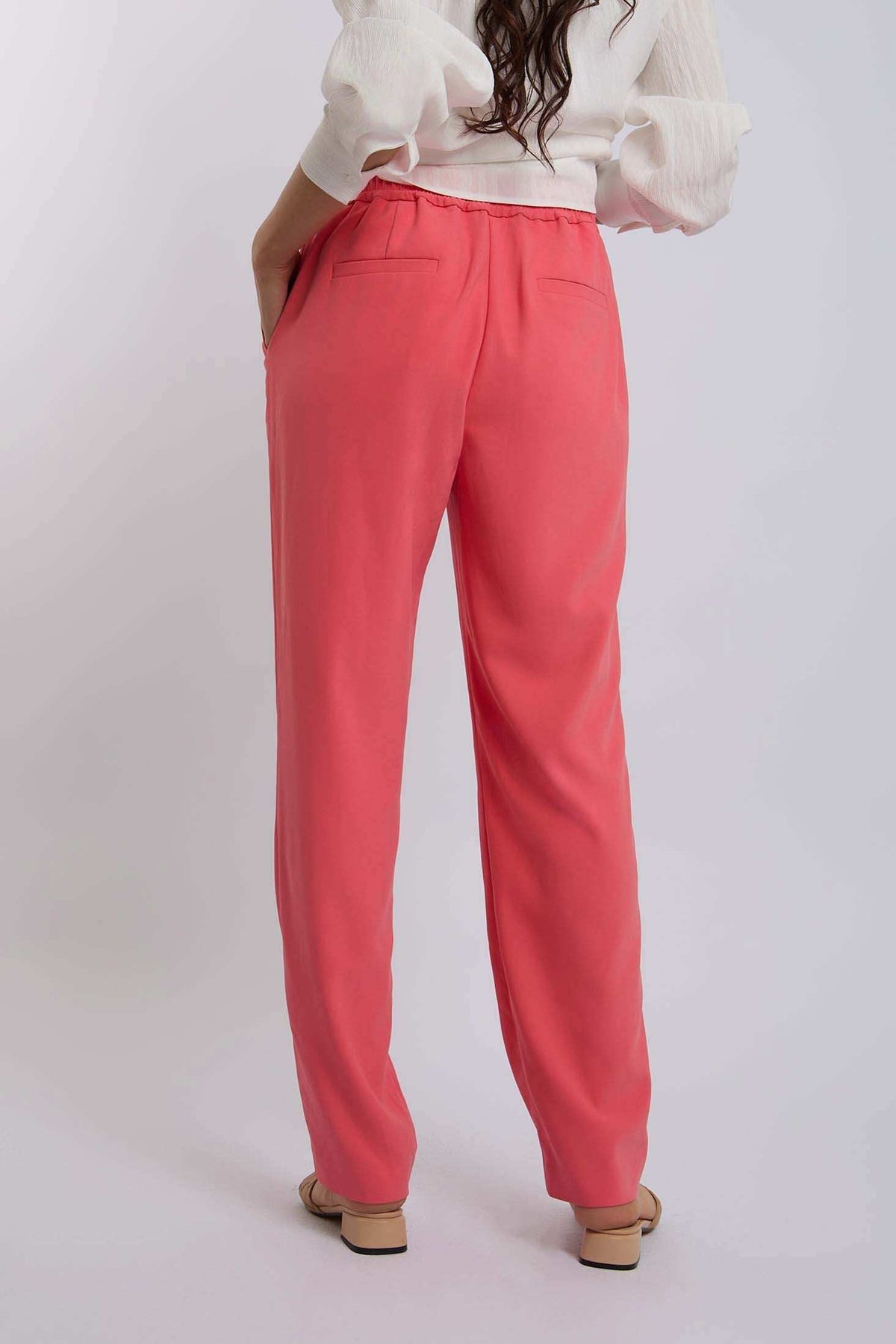 Woman in coral pink high-waisted pants with back pockets and white blouse, back view