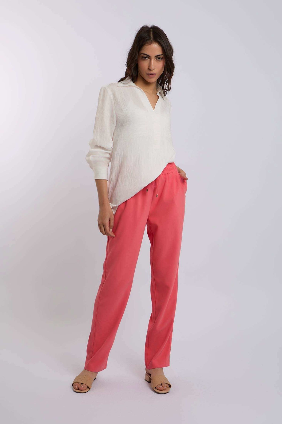 Woman wearing a white long sleeve blouse and coral pink pants, posing on a plain background