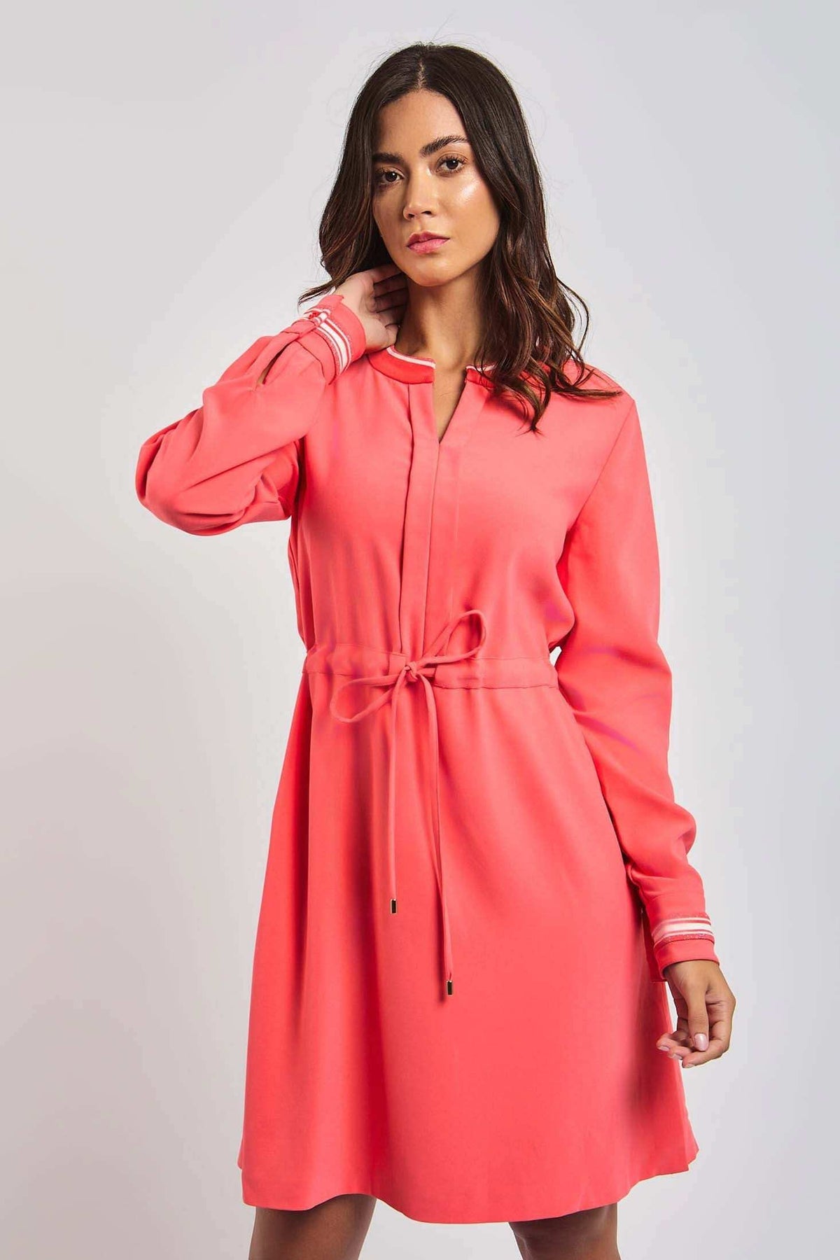 Woman in a coral pink long sleeve dress with waist tie and white cuff detail
