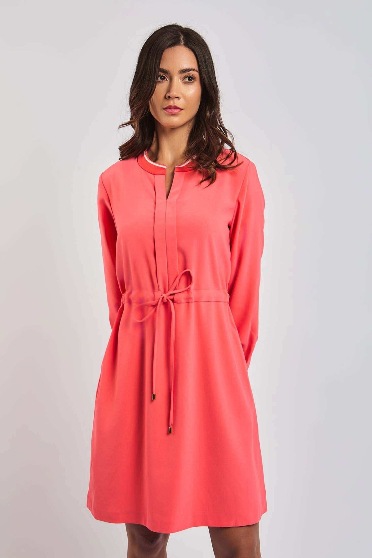 Woman wearing coral pink long sleeve casual dress with tie waist, standing against a plain background