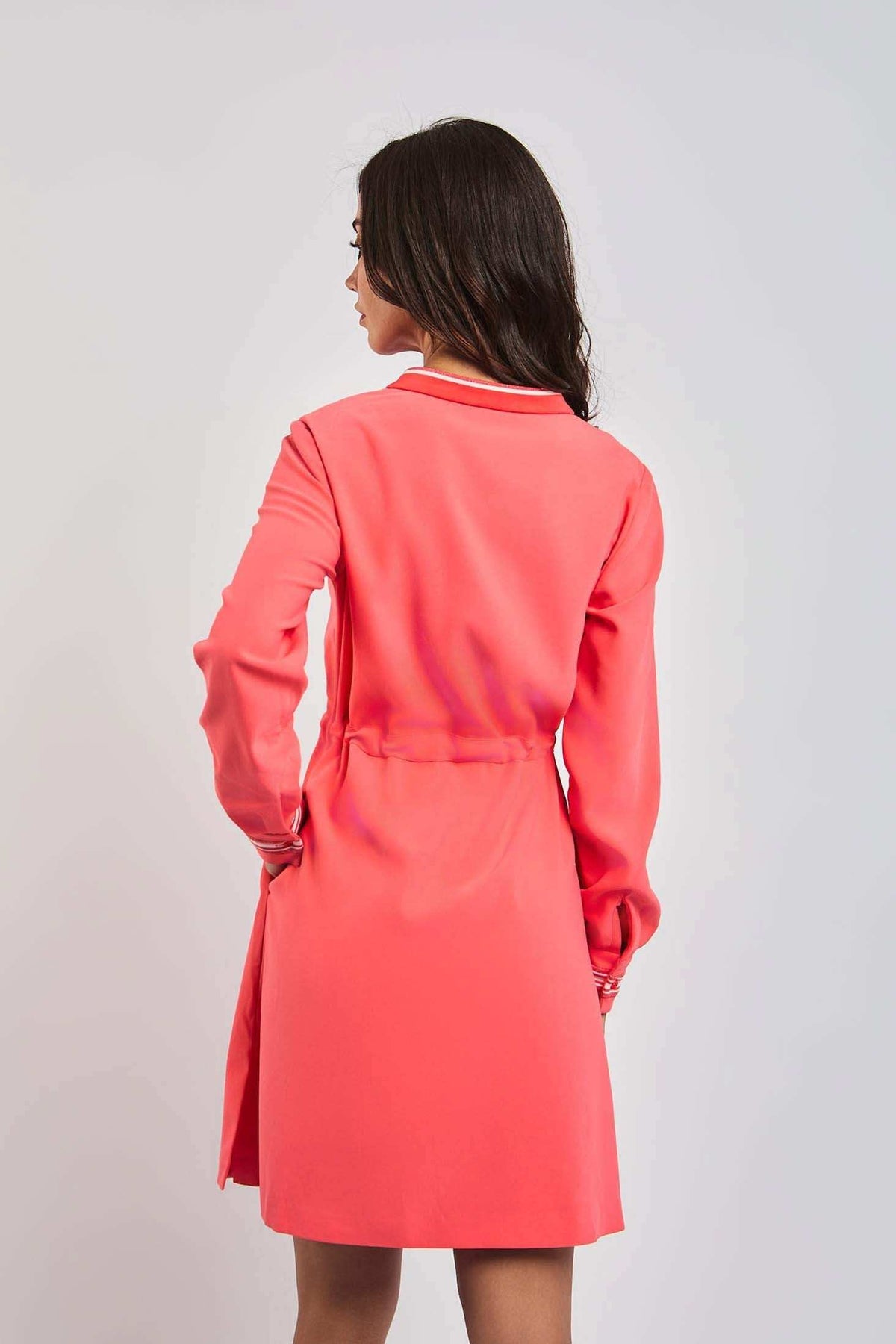 Woman wearing coral pink long sleeve dress, viewed from the back, against plain background